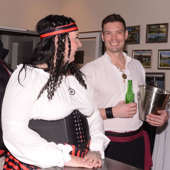 Eine Frau in einer Piratenkostüm spricht mit einem Mann im gleichen Kostüm, der eine grüne Flasche und einen Eimer hält. Sie lächeln und stehen neben einer Wand mit Bilderrahmen.