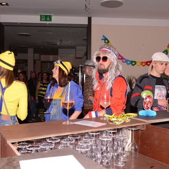 Mehrere Menschen in Kostümen stehen an einer Bar und halten Weingläser. Ein Mann hat einen langen Bart und trägt eine Perücke. Im Hintergrund schaut eine Frau in einem gelben Shirt den Mann an.