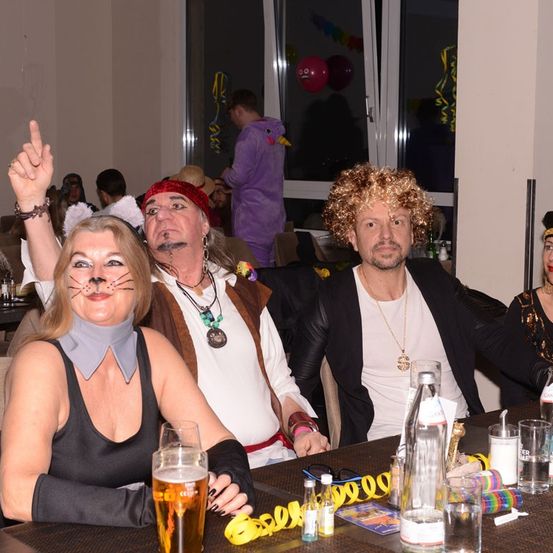 Vier Personen sitzen an einem Tisch und tragen Kostüme. Zwei Männer und zwei Frauen. Sie lächeln für die Kamera. Auf dem Tisch befinden sich Luftballons und Luftschlangen.