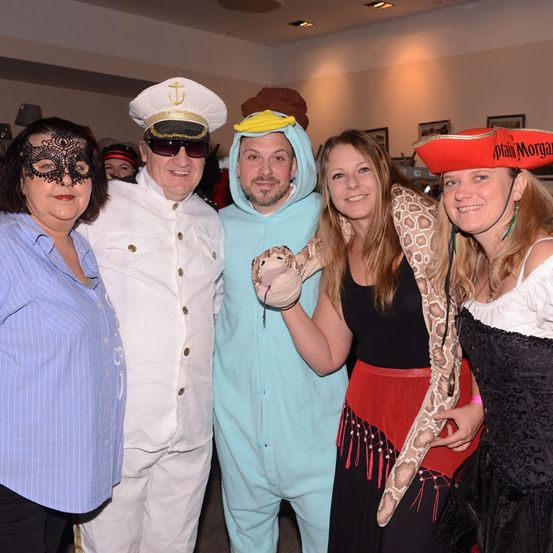 Fünf Menschen in Kostümen posieren für ein Foto. Ein Mann in Uniform und eine Frau in einem schwarzen Kleid halten eine Schlange. Die Frau in der Mitte trägt einen roten Rock und eine Maske. Hinter ihnen ist ein Raum mit weißen Wänden.