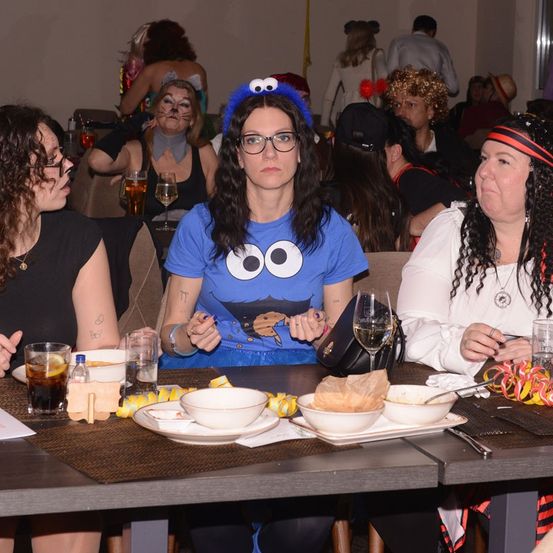 Mehrere Frauen sitzen in einem Saal an einem Tisch, einige in Kostümen. Eine Frau in einem blauen Shirt mit Augen isst. Es gibt Schüsseln, Weingläser und eine Tasche auf dem Tisch.