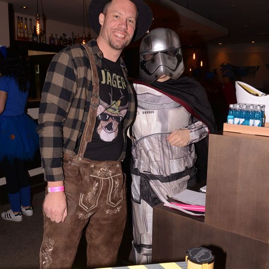 Ein Mann in traditioneller deutscher Kleidung steht neben einer Person in einem Star Wars Sturmtruppler-Kostüm bei einer Veranstaltung. Dahinter steht eine Frau in einem blauen Kleid und ein Tresen mit Flaschen.