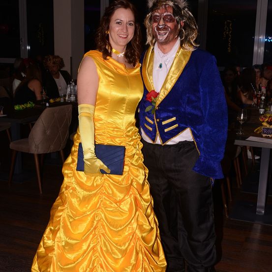 Eine Frau in einem gelben Ballkleid und ein Mann in einem blauen Kostüm mit weißem Make-up stehen zusammen, wahrscheinlich bei einer Veranstaltung. Dahinter sitzen mehrere Personen an Tischen mit Getränken.