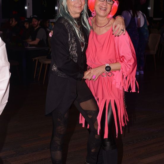 Zwei Frauen stehen bei einer Party nebeneinander und lächeln. Die Frau auf der linken Seite trägt ein schwarzes Kleid, während die Frau auf der rechten Seite ein rosa Kleid mit Fransen trägt. Hinter ihnen sitzen Leute an Tischen.