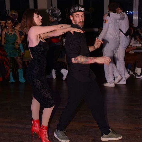 Ein Paar tanzt auf einer Party, beide in schwarzen Outfits. Die Frau hat rote Stiefel, der Mann Tattoos. Hinter ihnen stehen und sitzen andere Leute. Der Raum hat Holzböden und ist schwach beleuchtet.