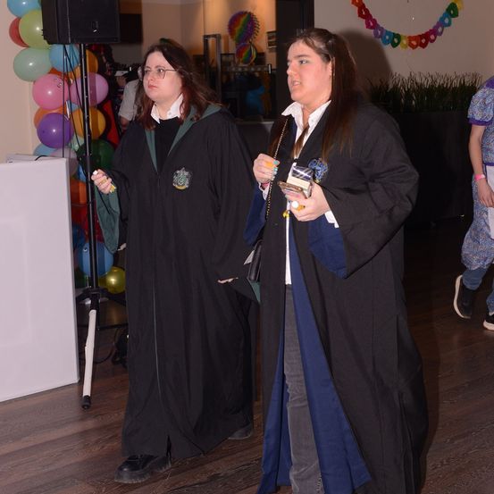 Zwei Frauen in Hogwarts-Roben gehen einen Flur mit Ballons, einem Lautsprecher und einer Pflanze entlang. Eine hält ein Handy.
