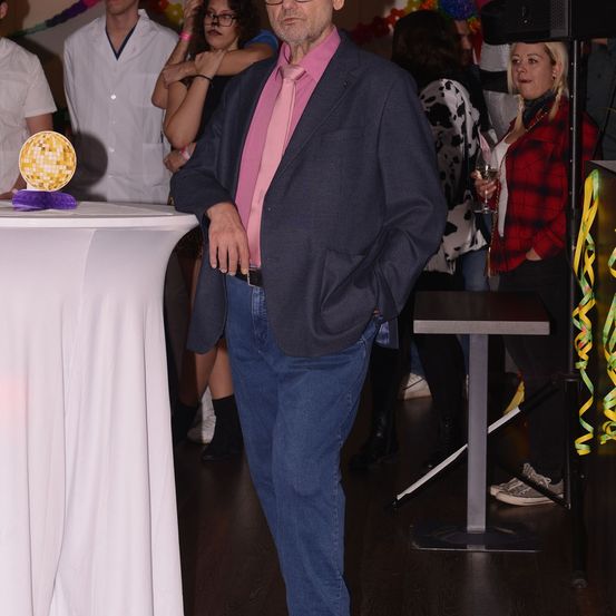 Ein Mann in einem rosa Hemd und Anzug steht in einer Partyumgebung, mit einer Discokugel und Luftballons im Hintergrund. Andere Leute, einschließlich einer Frau mit einem Getränk, sind ebenfalls anwesend.