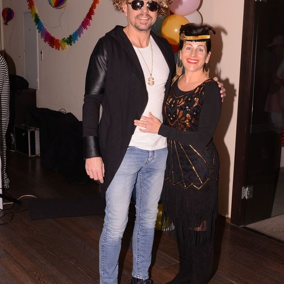 Ein Mann und eine Frau in schickem Kostüm posieren für ein Foto in einem Raum mit Luftballons und einer bunten Girlande.