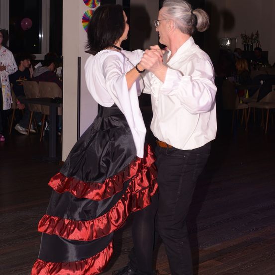 Ein älterer Mann und eine Frau tanzen in einem Raum. Die Frau trägt ein schwarz-rotes Kleid. Hinter ihnen sitzen Menschen auf Stühlen und Tischen. Ein Luftballon ist im Hintergrund.