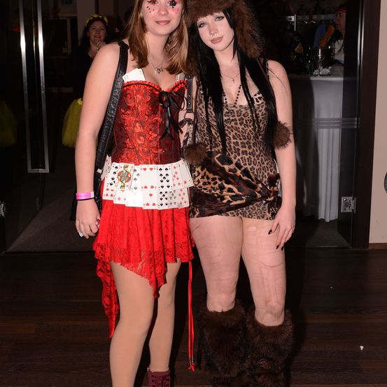 Zwei Frauen in Kostümen stehen zusammen in einem Raum mit dunklem Hintergrund. Eine trägt ein rotes Kostüm und die andere ein Leopardenmuster mit Pelzstiefeln.