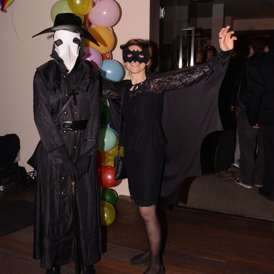 Ein Mann und eine Frau in schwarzen Kostümen stehen in einem Raum mit bunten Luftballons an der Wand. Der Mann trägt eine Maske.