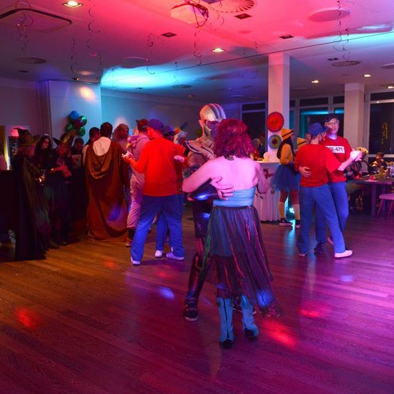 Eine Gruppe von Menschen in Kostümen tanzt auf einem Holzboden unter mehrfarbigen Lichtern. Eine Person mit Maske steht in der Mitte, umgeben von anderen.