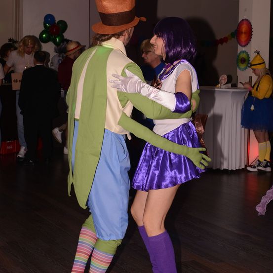 Ein Paar in Kostümen tanzt auf einer Party mit Luftballons und Menschen im Hintergrund.