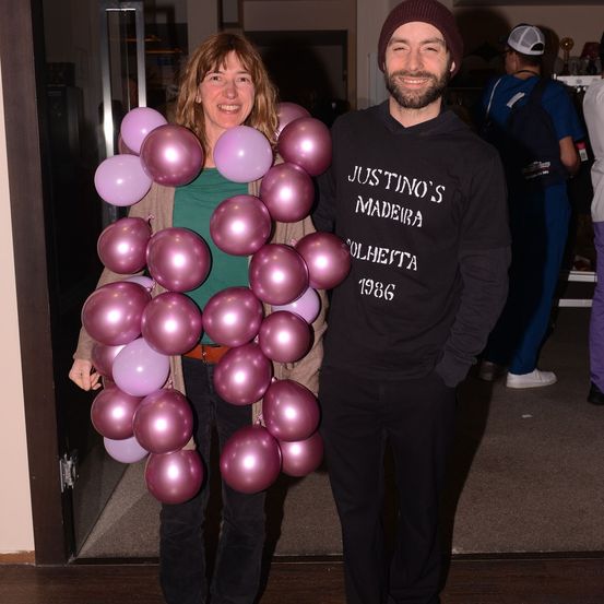 Eine Frau mit einem grünen Shirt hält eine Menge lila Luftballons. Sie steht neben einem Mann, der ein schwarzes Shirt mit den Worten 'Justino's Madeira Solheita 1986' trägt. Hinter ihnen steht ein Mann in Blau.