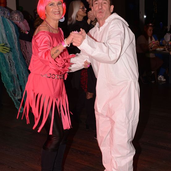 Ein Mann und eine Frau in rosa Kostümen tanzen in einer Partyumgebung, umgeben von anderen in festlicher Kleidung. Der Hintergrund ist schwach beleuchtet, mit mehr Menschen, die sitzen und essen.