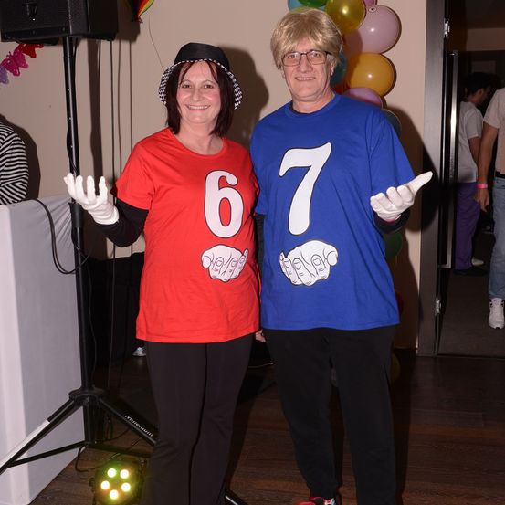 Zwei Erwachsene posieren für ein Foto. Die Frau trägt ein rotes Shirt mit der Nummer sechs. Der Mann trägt ein blaues Shirt mit der Nummer sieben. Hinter ihnen sind Luftballons und ein Lautsprecher.