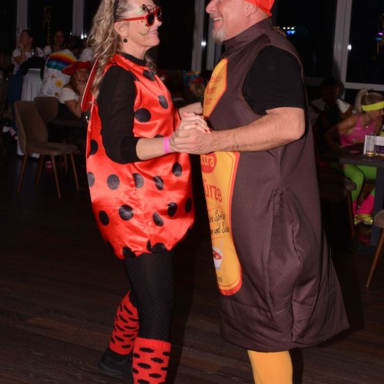 Ein Paar in Kostümen, möglicherweise für Halloween, tanzt auf einer Indoor-Party. Die Frau trägt ein rotes Marienkäfer-Kostüm, während der Mann ein braunes Soßenflaschen-Kostüm trägt.