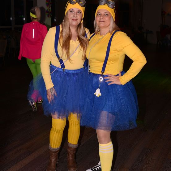 Zwei Frauen in Minion-Kostümen stehen zusammen. Beide tragen gelbe Oberteile und blaue Hosenträger. Die Frau auf der linken Seite hat ein Tutu und Stiefel. Im Hintergrund eine weitere Person in einem Kostüm.