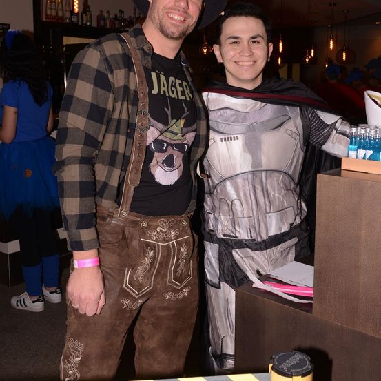 Zwei Männer stehen bei einer Party eng beieinander. Einer ist in einem schwarzen Shirt mit einem Hirsch gekleidet, der andere in einem silbernen und schwarzen Kostüm. Hinter ihnen befindet sich eine Bartheke mit Flaschen und einer Frau in einem blauen Kleid.