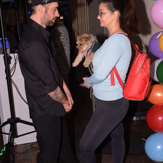 Ein Mann und eine Frau stehen sich gegenüber und halten Händchen. Die Frau hält einen kleinen Hund in ihren Armen. Auf ihrem Rücken befindet sich ein roter Rucksack. Hinter ihnen stehen viele bunte Luftballons.