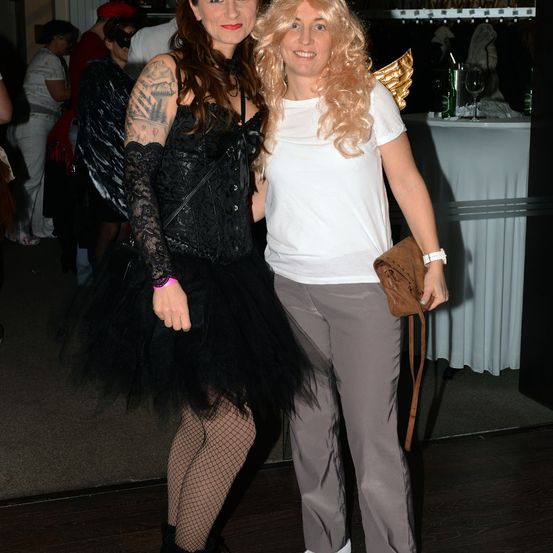 Zwei Frauen auf einer Party, eine in einem schwarzen Kostüm und die andere in einem weißen Shirt und grauen Hosen. Dahinter sind verschwommene Leute, ein Tisch und Regale.