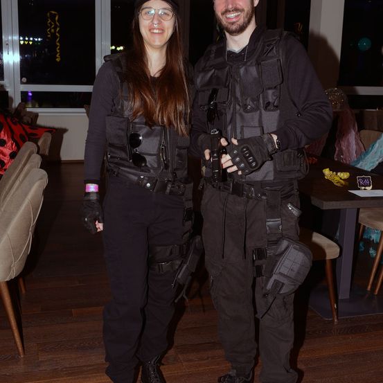 Zwei Personen in taktischer Ausrüstung, eine mit Brille und Hut, stehen in einer Innenumgebung. Sie haben Waffen, und dahinter stehen Stühle und ein Tisch.