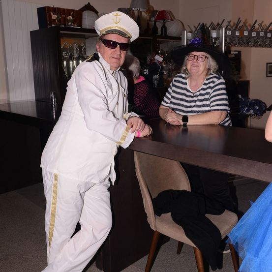 Ein Mann in einer Matrosenuniform steht neben einem Tisch, während eine Frau in einem gestreiften Shirt und Hut am Tisch sitzt. Es gibt einen Stuhl mit einem schwarzen Mantel darauf.