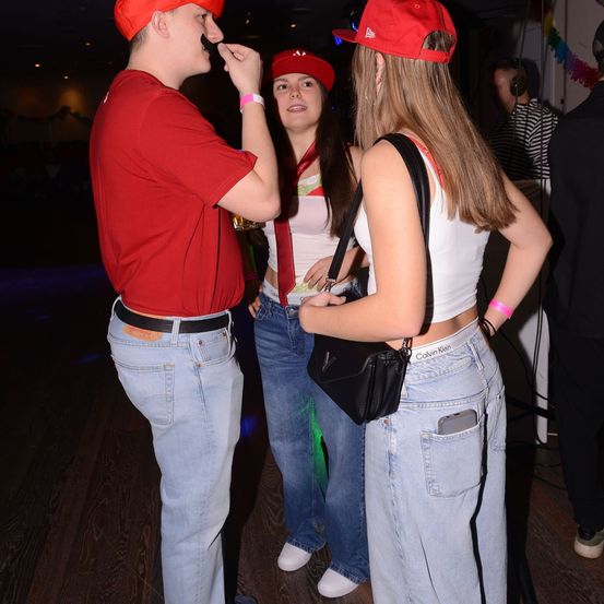 Drei junge Erwachsene, zwei Frauen und ein Mann, stehen in einem schwach beleuchteten Raum. Der Mann trägt ein rotes Shirt und eine rote Mütze, während die Frauen weiße und rosa Oberteile mit Jeans tragen. Sie scheinen zu unterhalten.
