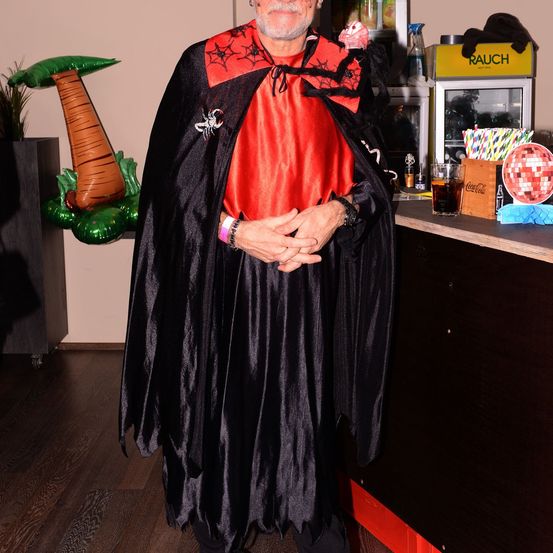 Ein Mann in einem roten und schwarzen Kostüm steht in einem Raum. Er hat eine Spinne auf der Brust. Hinter ihm steht eine Theke mit einer Coca-Cola-Box.