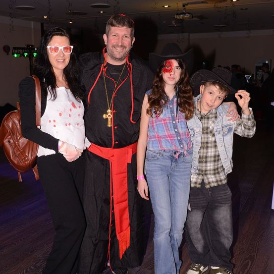 Vier Personen posieren für ein Foto. Der Mann ist als Priester gekleidet, während das Mädchen und der Junge Cowboyhüte tragen. Die Frau hat ein rot-weißes Oberteil. Sie stehen auf einem Holzboden.