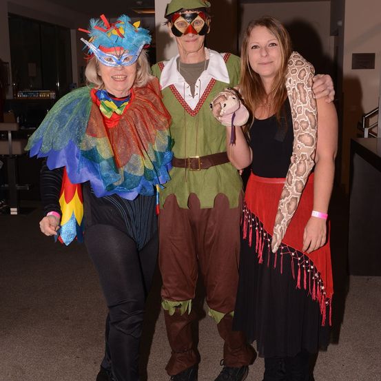 Drei Menschen in Kostümen posieren für ein Foto. Eine Frau in einem Federkostüm und einer Maske steht links. Ein Mann in einem grünen Kostüm steht in der Mitte. Eine Frau in einem roten Kleid hält eine Schlange rechts.