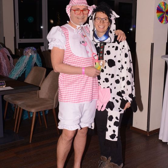 Zwei Erwachsene in Kostümen stehen eng beieinander in einem Raum und posieren lächelnd für ein Foto. Eine Person trägt ein Kuhkostüm.