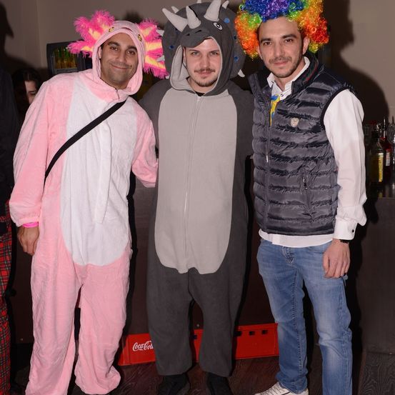 Drei Männer in Kostümen posieren für ein Foto. Einer trägt ein rosa Einhorn, ein anderer ein graues Dinosaurierkostüm und der dritte eine bunte Perücke.