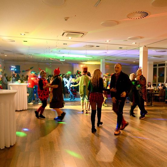 Eine Gruppe von Menschen tanzt in einem Raum mit bunten Lichtern und einem Holzboden. Einige tragen Kostüme, andere haben Ballons. Ein Tisch mit einer weißen Tischdecke steht in der Nähe.