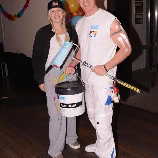 Ein Mann und eine Frau in bemalter Kleidung posieren mit Farbwalzen und einem Eimer Grundierung. Hinter ihnen schmücken Luftballons und eine festliche Girlande den Raum.
