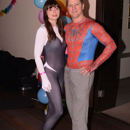 Ein Mann und eine Frau in Superheldenkostümen stehen nebeneinander in der Nähe einer Tür, mit bunten Luftballons im Hintergrund.