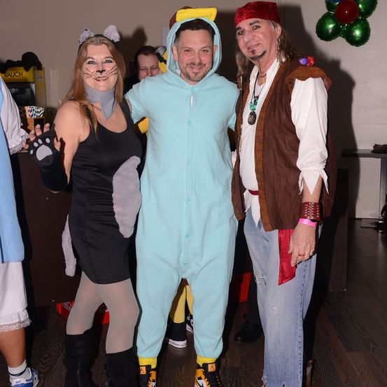 Drei Menschen in Kostümen posieren für ein Foto in einem Raum mit Holzboden und einer weißen Wand mit Luftballons. Eine Frau in einer Katzen-Kostüm, ein Mann in einem Babyanzug und ein Mann in einem Piratenkostüm.