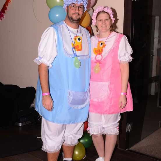 Ein Paar in Babykostümen mit Luftballondekoration im Hintergrund, der Mann in Blau und die Frau in Pink.