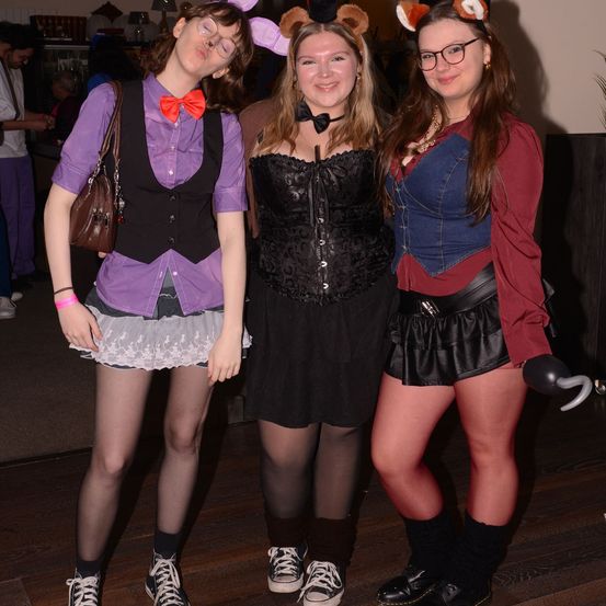 Drei Frauen in thematischen Kostümen posieren für ein Foto. Die Frau auf der linken Seite trägt ein lila Hemd und eine schwarze Weste, die mittlere ein schwarzes Korsett und die rechte ein rotes Hemd und eine Jeansweste. Alle tragen Kopfbedeckungen mit Tierohren.