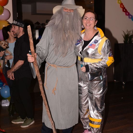 Ein Mann als Gandalf verkleidet und eine Frau in einem silbernen Raumanzug stehen bei einer Veranstaltung zusammen. Hinter ihnen halten zwei Personen einen Hund und eine weitere Person steht. Im Hintergrund befinden sich Luftballons.