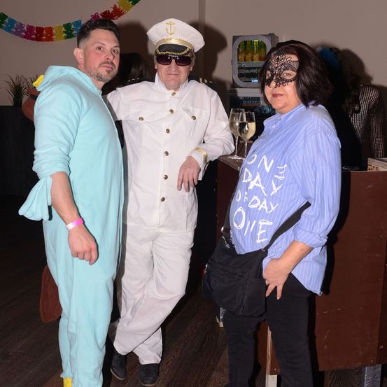 Drei Erwachsene stehen bei einer Party. Ein Mann in Blau und Gelb, ein Mann in einem Kapitänsuniform und eine Frau in einer schwarzen Maske und einem blauen Shirt. Dahinter steht eine Theke mit zwei Gläsern Wein.