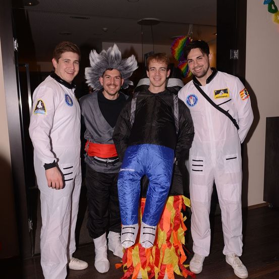 Vier Männer in Kostümen stehen zusammen, einer mit einem Flammendesign, ein anderer mit einer grauen Perücke und die anderen beiden in Astronautenanzügen. Sie lächeln für ein Foto.