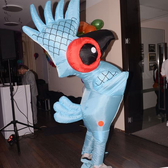 Eine Person in einem blauen Vogelkostüm steht drinnen. Es gibt Luftballons und eine Person im Hintergrund.