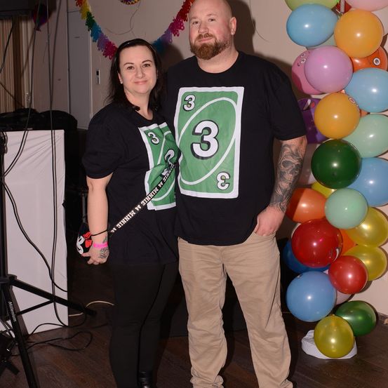 Ein Mann und eine Frau stehen zusammen vor einem farbenfrohen Ballonbogen. Der Mann trägt ein schwarzes T-Shirt mit einer Nummer-drei-Grafik, während die Frau einen Schläger hält.