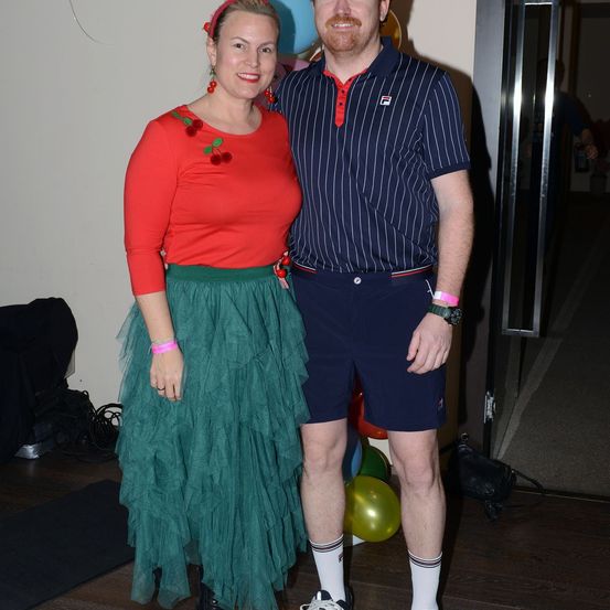 Ein Mann und eine Frau stehen eng beieinander in einem Raum und tragen festliche Kleidung. Die Frau hat einen roten Pullover und einen grünen Rock, während der Mann ein blaues Polohemd und Shorts trägt. Sie sind von Luftballons umgeben.