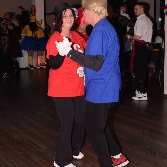 Eine Frau in einem roten Shirt tanzt mit einem Mann in einem blauen Shirt auf einer Party mit anderen in Kostümen.