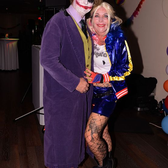 Ein Paar, gekleidet als Joker und Harley Quinn, steht nah beieinander und lächelt für die Kamera. Sie sind in einem Raum mit Luftballons und einem Tisch im Hintergrund.