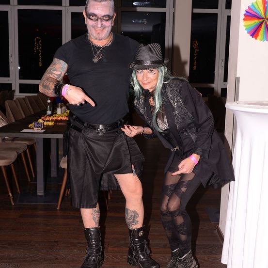 Ein Mann und eine Frau posieren für ein Foto, beide in schwarzen Outfits. Der Mann hat Gesichtsbemalung und einen Kilt, während die Frau einen Hut und Strumpfhosen mit Spitze trägt. Sie stehen neben einem Tisch mit verschiedenen Gegenständen.