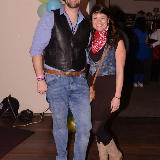 Ein Paar in Western-Kleidung steht nah beieinander und lächelt, mit Luftballons und einer Lampe im Hintergrund.