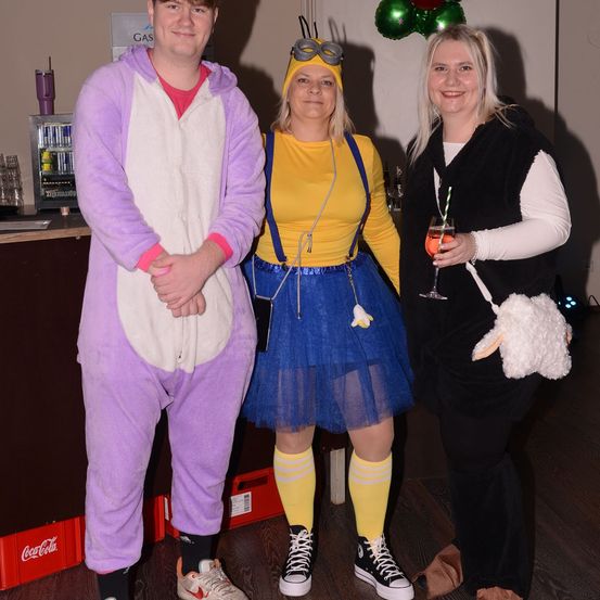 Drei Menschen in Kostümen stehen zusammen. Ein Mann in einem lila Bärenkostüm, eine Frau in einem gelben Minion-Kostüm und eine weitere Frau in einem schwarzen und weißen Kostüm. Sie lächeln für ein Foto.
